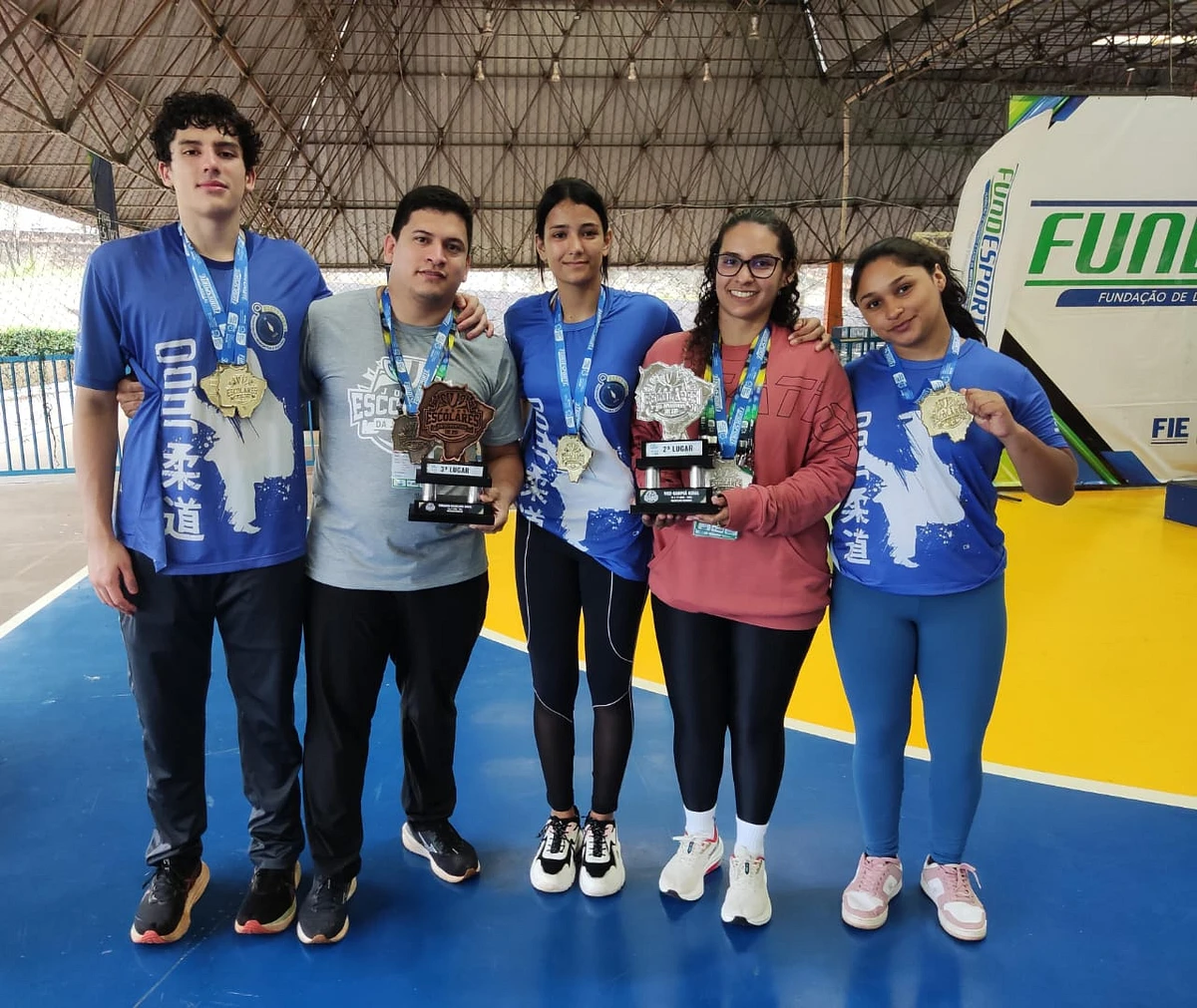 Os técnicos Edimir Kenedi e Ana Pedro exibem os troféus conquistados por São Gabriel do Oeste nos Jogos Escolares, ao lado dos atletas medalhistas do Wrestling.
