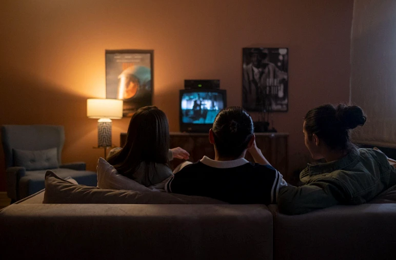 Três pessoas assistindo televisão juntas no sofá em sala aconchegante com iluminação quente e pôsteres de filmes na parede.